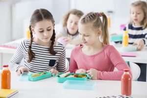 Two young girls eating free school meals and looking into each other's lunch boxes with healthy vegetables and bread. Bottles of fruit juices on the desk. Other kids in blurred background.